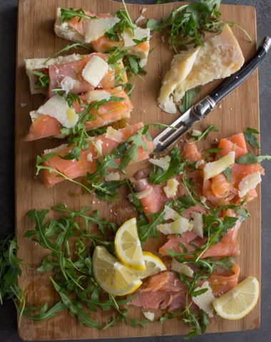 smoked salmon slices, parmesan cheese chunks, rucola leaves, lemon slices and a knife on a wooden board